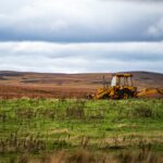 A solitary yellow backhoe on a wide green field against a backdrop of hills and a cloudy sky.